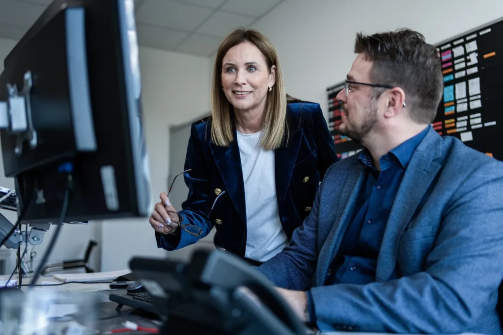 Ein Mann sitzt an einem Schreibtisch. Eine Frau steht hinter ihm und sie schauen auf den Bildschirm