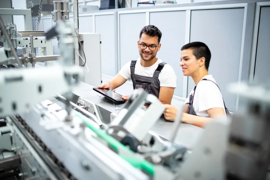 Zwei Menschen in Latzhose halten ein Tablet und schauen auf eine Maschine